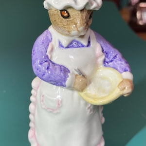 Op de afbeelding: Een keramieken beeldje van een konijn met een witte muts, een paars shirt met lange mouwen en een wit schort met roze ruches. Het konijn houdt een gele mengkom vast met beide handen. De voet van het beeldje is grijs geschilderd.