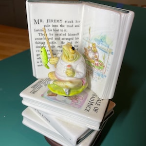 May include: A ceramic figurine of Mr. Jeremy Fisher sitting on a lily pad, holding a fishing pole with a green fish. The figure is placed in front of an open book page with text and illustrations, resting on a stack of two white ceramic books.