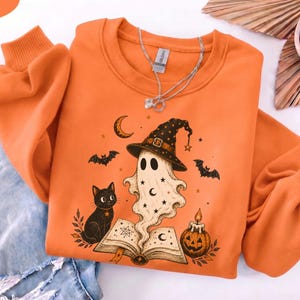 May include: An orange crewneck sweatshirt featuring a graphic print of a ghost wearing a witch hat, a black cat, an open book, a pumpkin, bats, and stars. The sweatshirt has long sleeves and a ribbed collar, with a silver-toned necklace resting on top.