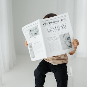 Op de afbeelding: Een kind zit op een doorzichtige stoel en houdt een krant vast met de titel "The Birthday Post". De kop luidt "Levi's First Birthday". Het kind draagt een beige shirt en een zwarte broek. De krant heeft een vintage uitstraling.