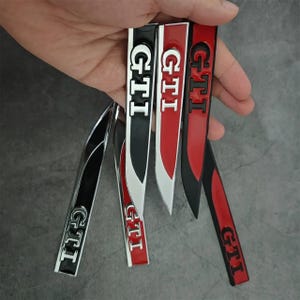 Pode incluir: Uma coleção de emblemas de carro GTI em vários esquemas de cores, incluindo preto, vermelho e prata. Os emblemas são alongados e apresentam o logotipo GTI em uma fonte branca. O fundo é cinza neutro.