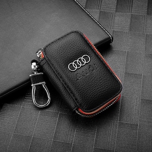 May include: Black leather key case with the Audi logo and the name "Audi" embossed on the front. The case has a red and white zipper and a silver key ring attachment.