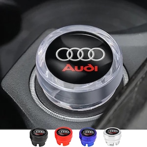 May include: Clear, circular car accessory featuring the Audi logo in white and red. The accessory is set in a car's console. Below are four additional accessories in black, red, blue, and silver.