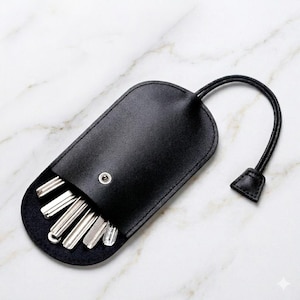 May include: A black leather key holder with a rounded rectangular shape and a silver snap button closure. The case contains several silver metal keys protruding from the bottom, and it features a thin black leather strap with a small tab.