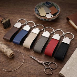 以下が含まれることがあります： シルバー調の金属製金具が付いた6つのレザーキーホルダーが並んでいます。色はダークブラウン、ブルー、ベージュ、グレー、レッド、ブラックです。小さなボウルには様々な金属製のタグが入っており、近くにはハサミが置かれています。