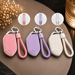 May include: Three car key fob covers in pink, lavender, and white, each featuring a matching braided leather strap and a metal keychain. The covers have a smooth, glossy finish and include a side view inset showing the button layout for each colour.