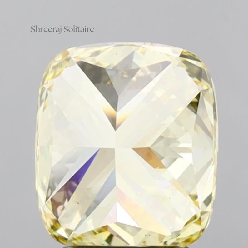 May include: A cushion-cut diamond with a pale yellow hue. The gemstone has numerous facets that reflect light, creating a sparkling effect. The text "Shreeraj Solitaire" is in the upper left corner.