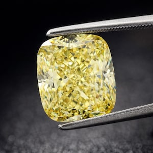 May include: A cushion-cut, fancy yellow diamond held by tweezers against a black background. The diamond's facets reflect light, creating a brilliant sparkle. The shape is square with rounded corners, showcasing its unique cut.