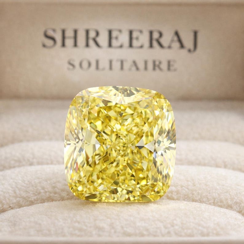 May include: A large, cushion-cut, fancy vivid yellow diamond. The diamond is set in a neutral-toned box with the words "SHREERAJ SOLITAIRE" printed in a dark font. The diamond's facets reflect light, creating a brilliant display.