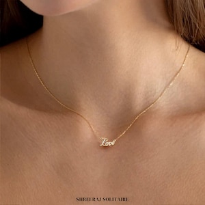 Puede incluir: Un collar de cadena de oro con un delicado colgante que dice "Love" en letra cursiva. El collar lo lleva una persona de tez clara.