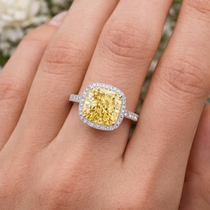 May include: A square-cut yellow gemstone ring with a halo of smaller white gemstones. The ring's band is also adorned with small white gemstones. The ring is set on a hand, showcasing the ring's design and sparkle.