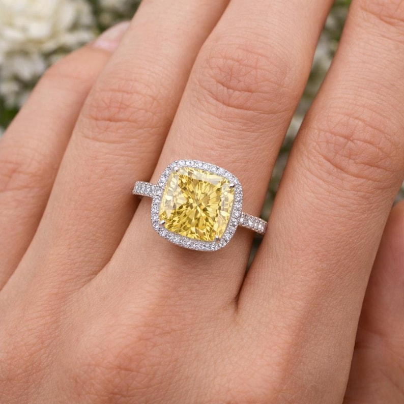 May include: A square-cut yellow gemstone ring with a halo of smaller white gemstones. The ring's band is also adorned with small white gemstones. The ring is set on a hand, showcasing the ring's design and sparkle.