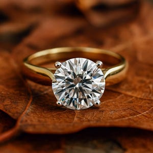 May include: A gold engagement ring featuring a large, round, clear gemstone. The ring is set against a brown leaf, contrasting the shiny ring with the natural background. The ring's band is smooth and polished.
