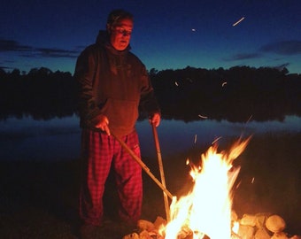 May include: A man wearing a brown hoodie and red checked trousers stands by a campfire at night. He is holding two sticks over the fire.