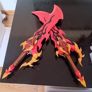 Może przedstawiać: Dwa skrzyżowane czerwono-złote miecze fantasy o skomplikowanych wzorach. Ostrza mają wzór płomienia w kolorze czarnym, a rękojeści są brązowe ze złotymi akcentami. Miecze są prezentowane na czarnej powierzchni.