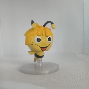 May include: A figurine of a cartoon bee with yellow hair, black antennae, and a black and yellow striped body. The bee has a smiling face with large eyes and is mounted on a clear, circular base.