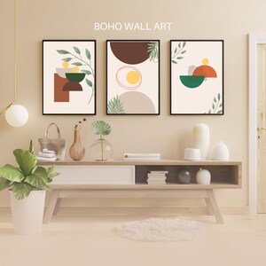 Puede incluir: Tres impresiones de arte de pared abstracto enmarcadas con formas geométricas y follaje en tonos de marrón, verde, naranja y amarillo. Las impresiones están colgadas en una pared beige sobre una mesa consola de madera.