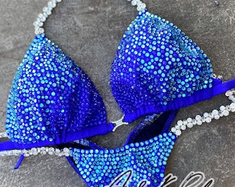 May include: A blue bikini with sparkling embellishments. The top and bottom are covered in small, iridescent stones that transition from light to dark blue. The bikini has a halter top design with rhinestone accents and the text "A&K Bikini".