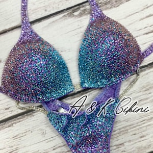 Puede incluir: Un bikini de lentejuelas morado y azul con un top halter y una parte inferior de corte alto. El bikini está decorado con pedrería y tiene un diseño degradado. El texto "A & K Bikini" es visible en la parte inferior.