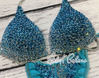 May include: A turquoise blue bikini with crystal embellishments. The bikini top has a halter style with a silver chain strap. The bikini bottom has a high-cut style with a ruffle detail. The bikini is decorated with a gradient of blue crystals.