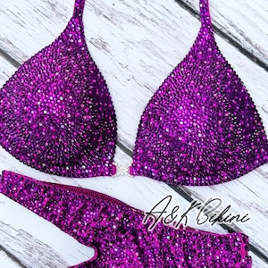 Puede incluir: Un bikini morado con un diseño brillante, similar a cristales. La parte superior del bikini tiene forma triangular y la parte inferior es de estilo de corte alto. El bikini está sobre una superficie de madera blanca. El texto "A&K Bikini" es visible en la imagen.