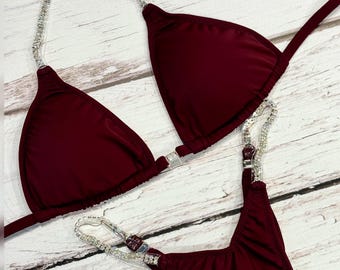 May include: A burgundy bikini set with rhinestone accents. The top is a triangle style, and the bottoms are a thong cut. The straps and edges are embellished with sparkling rhinestones. The image includes the text "A&K Bikini Shop".