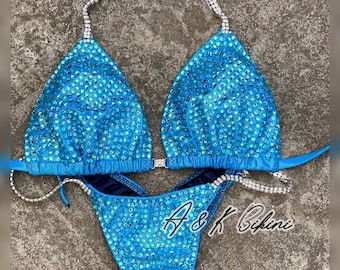 May include: A turquoise blue bikini with crystal embellishments. The bikini top has a triangle shape and the bottom is a high-cut style. The bikini is adorned with silver rhinestones and has a silver chain strap.