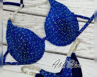 May include: A blue and silver competition bikini with crystal embellishments. The bikini top has a halter style strap and the bottom is a V-shaped cut. The bikini is adorned with rhinestones and crystals.