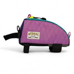 May include: A colourful bicycle handlebar bag with a purple diamond-patterned front. The bag has teal, purple, and yellow accents, with black straps and the word "STICAL" on a patch.