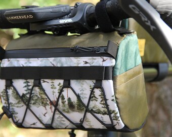 Pode incluir: Uma bolsa de guiador verde e castanho com um estampado de floresta branco e verde. A bolsa tem um fecho de correr e uma correia preta com uma fivela. A bolsa está presa ao guiador de uma bicicleta.