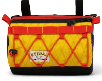 May include: A multi-colored bag with a yellow front, red accents, and black straps. The bag features a diamond pattern on the yellow section and the word "STICAL" on a white patch. The bag has a zipper closure and red bungee cords.