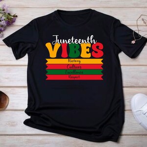 May include: Black t-shirt with the text "Juneteenth Vibes" in colorful letters. Below the text are four horizontal stripes in red, yellow, green, and red, with the words "History", "Culture", "Excellence", and "Respect" printed on each stripe.