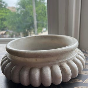 May include: A white marble decorative bowl with a wide, fluted base and a smooth, rounded rim. The bowl has a classic design and is suitable for various decorative purposes. The bowl is made of a light-colored stone.