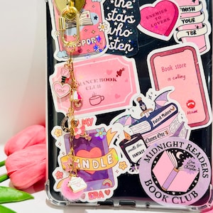May include: A phone case covered in pink and white stickers with book-related themes, such as "Midnight Readers Book Club" and "Enemies to Lovers." A gold charm with a heart and a pink pom-pom hangs from the case. The overall aesthetic is centered around books and reading.