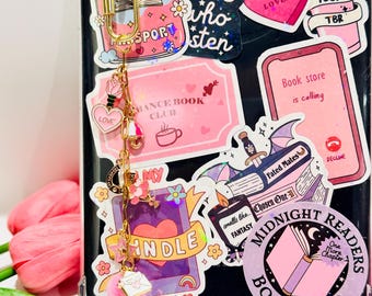Könnte beinhalten: Eine transparente Handyhülle mit verschiedenen rosa und weißen Aufklebern, darunter "Midnight Readers Book Club" und "Enemies to Lovers". Ein goldener Charm mit Herz und rosa Bommel hängt an der Hülle. Das Thema dreht sich um Bücher und Lesen.