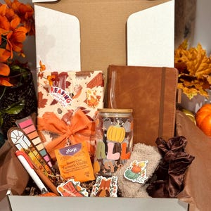 May include: A gift box filled with fall-themed items. Includes a brown leather journal, a glass tumbler with a pumpkin design, a book, pens, stickers, tea, and socks. The box is decorated with orange and brown accents.