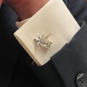 Può includere: Gemelli a forma di tartaruga argentata su un polsino di camicia color crema. I gemelli presentano dettagli di pinne e guscio. La camicia è abbinata a una giacca da abito blu scuro.