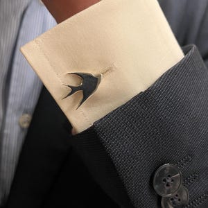 May include: Silver-toned swallow cufflinks on a white dress shirt cuff, paired with a dark gray suit jacket. The cufflinks feature a detailed bird design, adding a touch of elegance to formal attire. The jacket has three dark buttons.