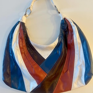 May include: A vintage shoulder bag with a patchwork design. The bag features alternating vertical stripes of white, navy blue, brown, and red leather. It has a white strap and silver metal rings.