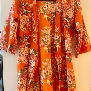 May include: An orange floral kimono-style robe with a vibrant rose pattern. The robe is hanging on a black hanger against a white wall. The floral design includes shades of pink, white, and green.