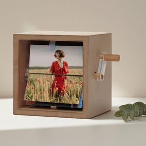 Könnte beinhalten: Ein hölzerner Fotowürfel mit einem Griff aus klarem Acryl und Bambus. Der Würfel zeigt ein Foto einer Person in einem roten Kleid in einem Feld roter Blumen. Der Würfel ist für die Aufnahme und Anzeige mehrerer Fotos konzipiert.