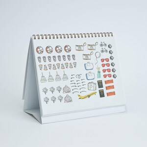 May include: A white desk calendar with a spiral binding, featuring colorful illustrations of travel-related items. The drawings include cameras, suitcases, sunglasses, pizza slices, and a yellow airplane. The calendar is open and angled, displaying the current month's designs.