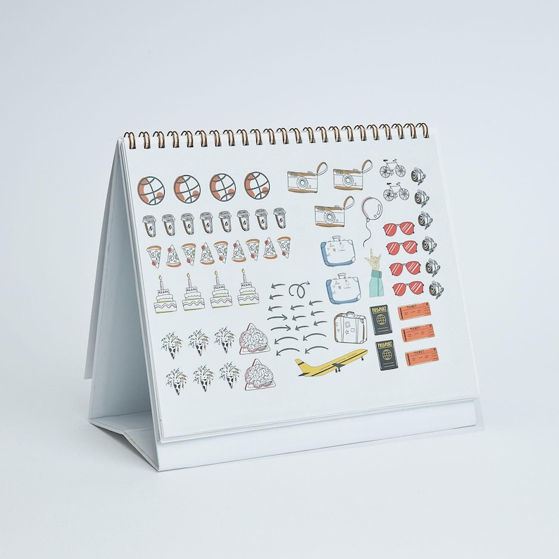 May include: A white desk calendar with a spiral binding, featuring colorful illustrations of travel-related items. The drawings include cameras, suitcases, sunglasses, pizza slices, and a yellow airplane. The calendar is open and angled, displaying the current month's designs.