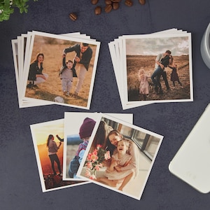 May include: A collection of square photo prints featuring family portraits. The images showcase various family moments, including outdoor scenes with parents and children. The prints are arranged on a dark surface, with coffee beans and a smartphone nearby.