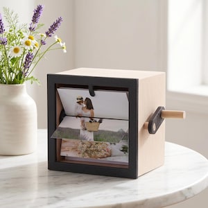 May include: A wooden photo cube with a black frame and a wooden handle. The cube displays three photos, each partially visible. The cube is sitting on a white marble table. A vase of flowers is in the background.