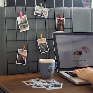K&ouml;nnte beinhalten: Ein schwarzes Drahtgitter mit mehreren Fotos, die mit farbigen W&auml;scheklammern befestigt sind. Ein blauer Becher mit Kaffeemotiv steht auf einem Holztisch, zusammen mit einem Laptop und mehreren losen Fotos. Der Laptop-Bildschirm zeigt eine Website.