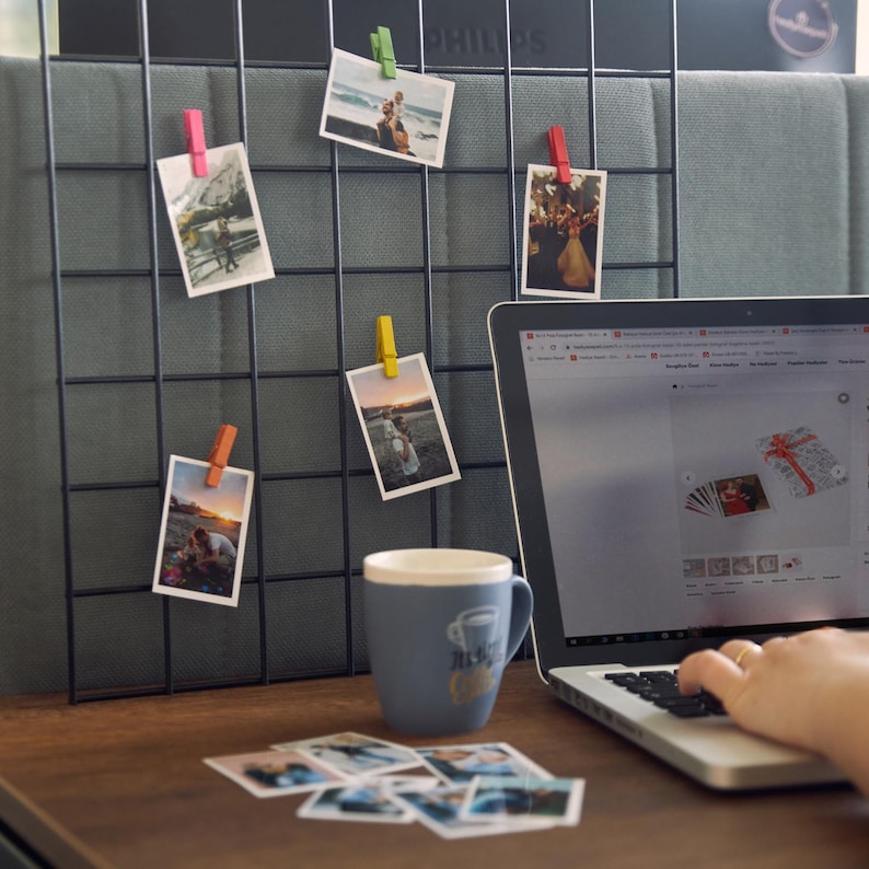 K&ouml;nnte beinhalten: Ein schwarzes Drahtgitter mit mehreren Fotos, die mit farbigen W&auml;scheklammern befestigt sind. Ein blauer Becher mit Kaffeemotiv steht auf einem Holztisch, zusammen mit einem Laptop und mehreren losen Fotos. Der Laptop-Bildschirm zeigt eine Website.
