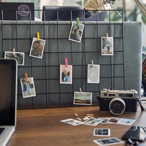 K&ouml;nnte beinhalten: Ein schwarzes Drahtgitter-Fotodisplay mit mehreren kleinen Fotos, die mit farbigen W&auml;scheklammern befestigt sind. Eine Vintage-Kamera und weitere Fotos befinden sich auf dem Holzschreibtisch. Die Fotos scheinen Familienfotos zu sein.