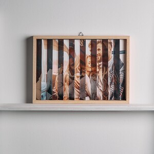 May include: A framed photo print with a light wood frame. The image is segmented into vertical strips, revealing a collage of people. The photo is displayed on a white shelf against a white wall.