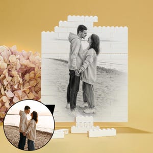 May include: A pencil sketch of a couple holding hands on a beach is displayed on a white brick wall. The wall is constructed from large white building blocks. A smaller, circular image of the couple is in the lower left corner. A bouquet of pink flowers is on the left.